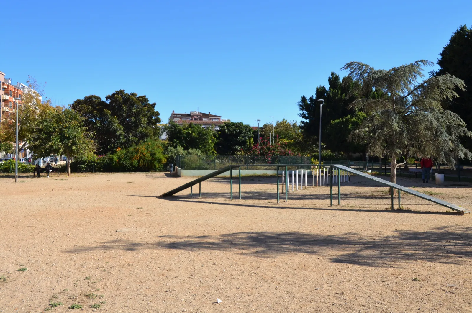 Sandy dog exercise area in a park with a wooden ramp, trees and apartment blocks behind
