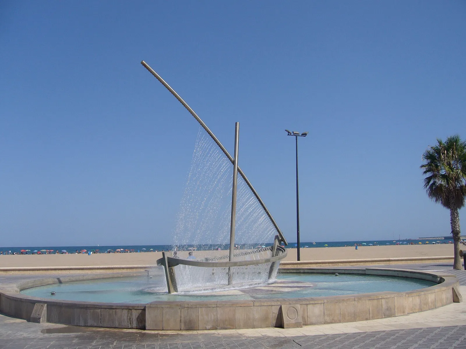 Fountain shaped like a sail with water falling into a round basin beside a sandy beach