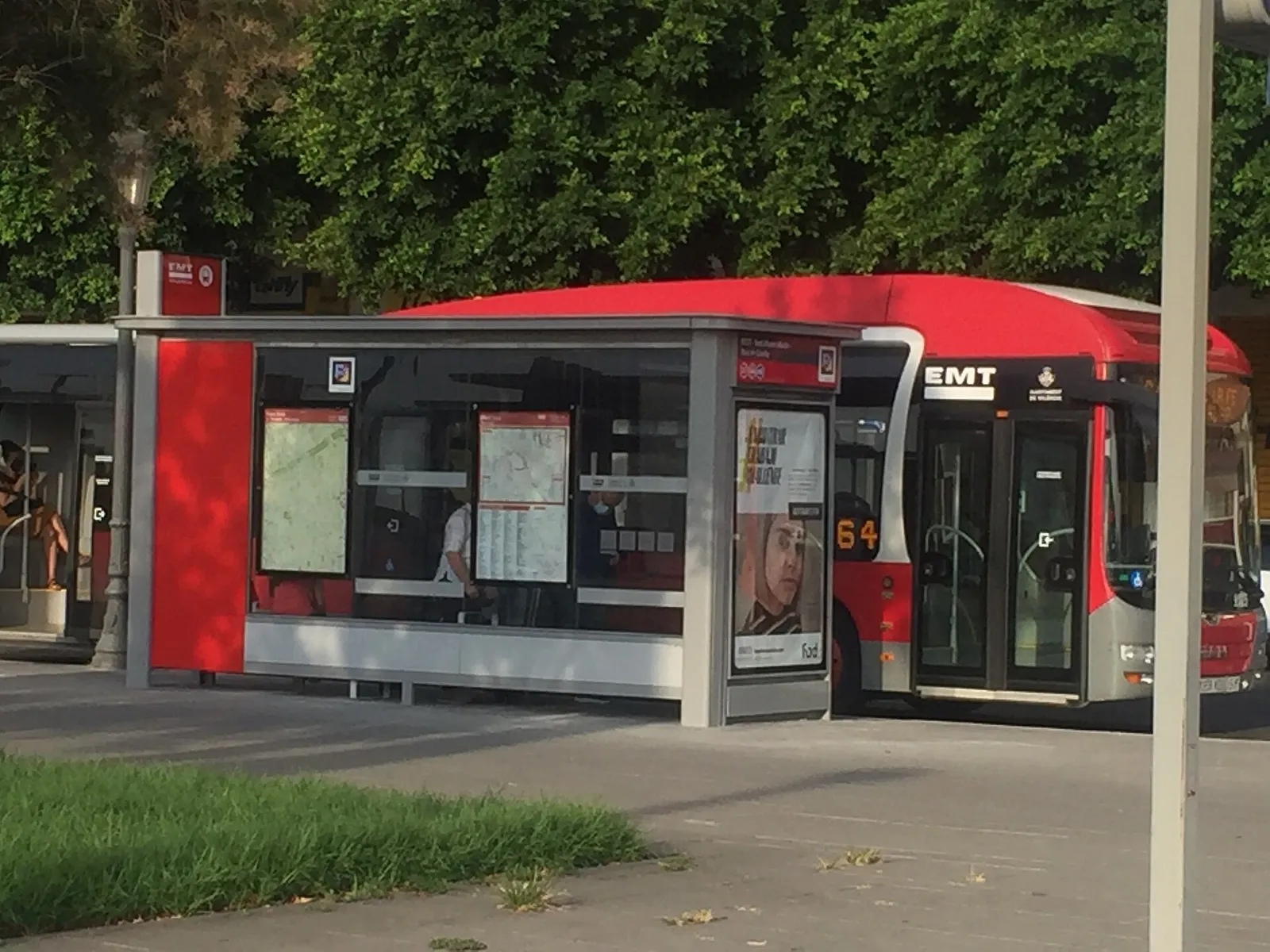 Red city bus beside a covered bus stop with a timetable panel and trees behind