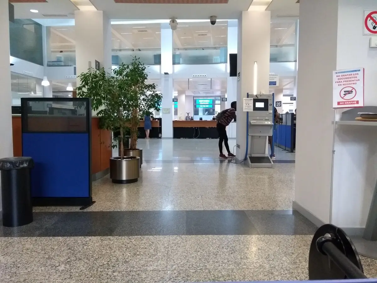 Public hall inside a tax office with a service counter, a plant and a floor of speckled stone.