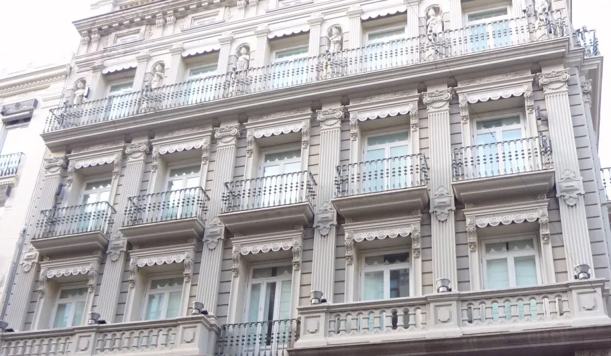 Pale stone residential building with balconies and ground-floor shop windows on a city street.