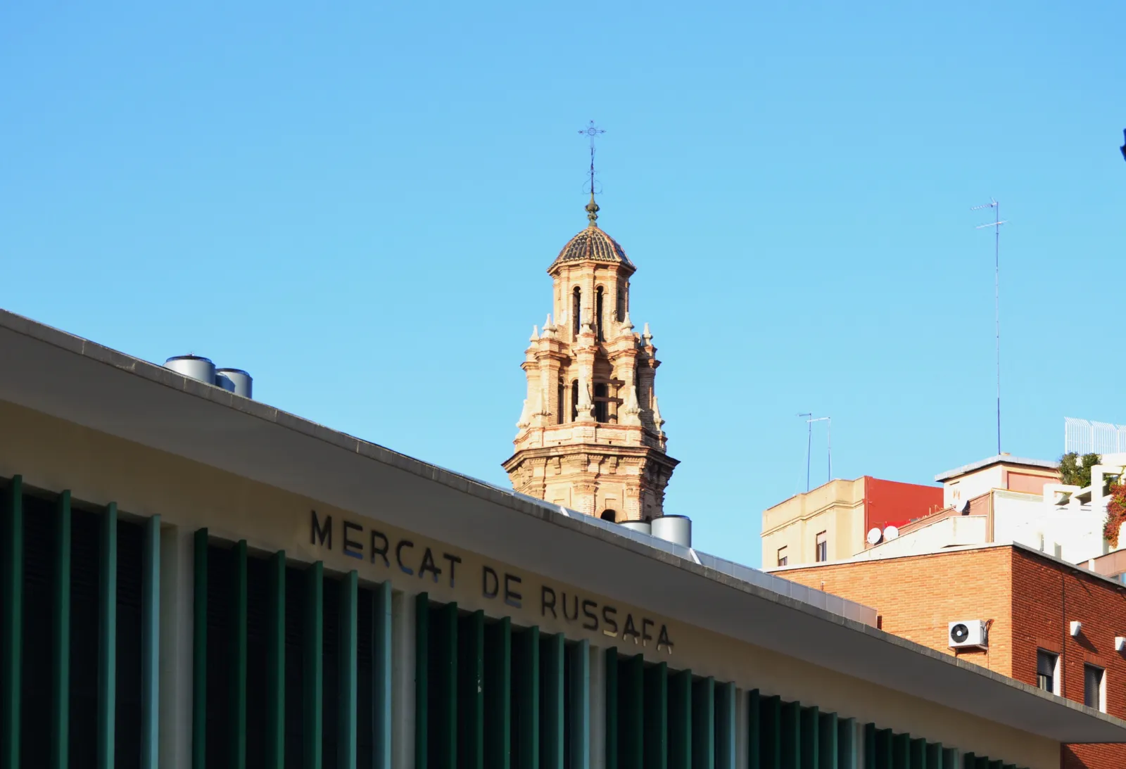 Roof edge of the Mercat de Russafa with its name on the facade and a church bell tower behind.