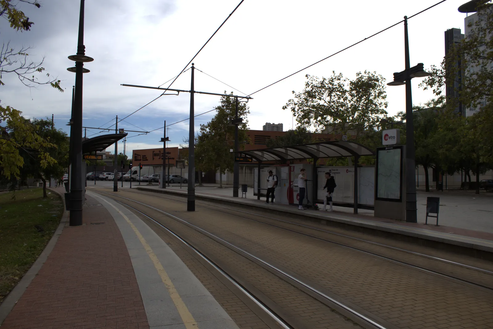 Covered tram stop beside a tree-lined track with passengers waiting on the platform