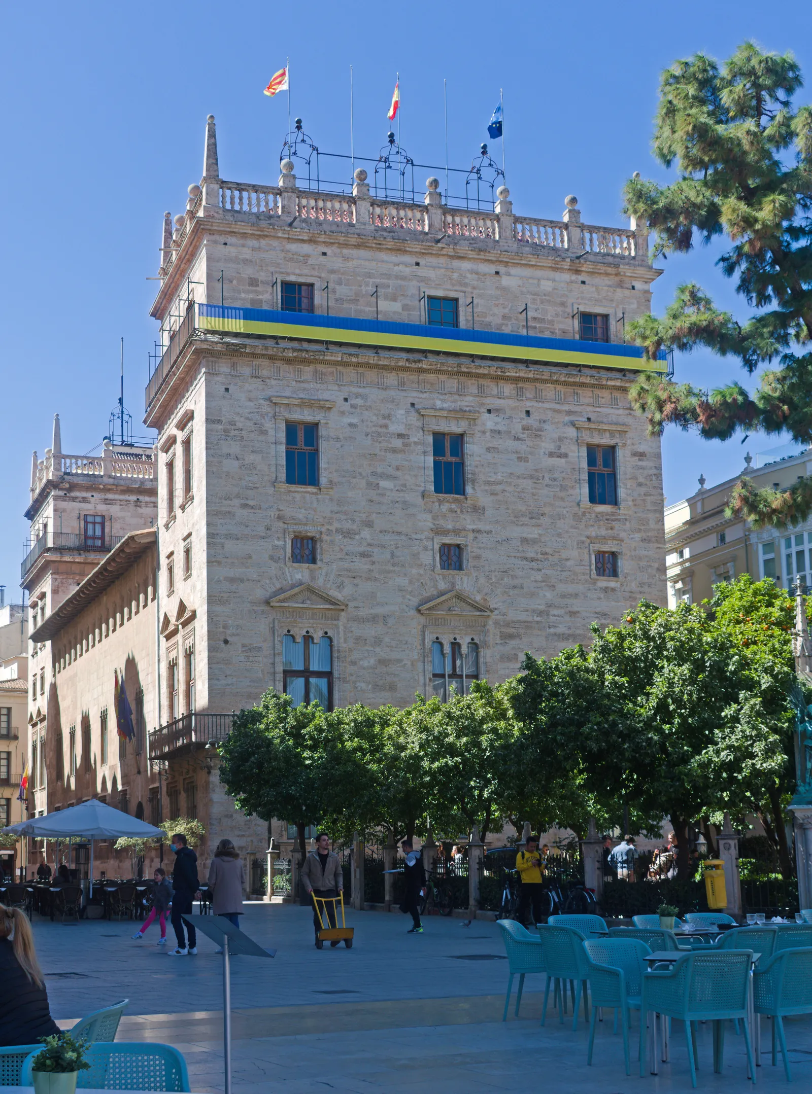 Historic palace with a stone tower and rows of arched windows, with a street cafe and trees in front.