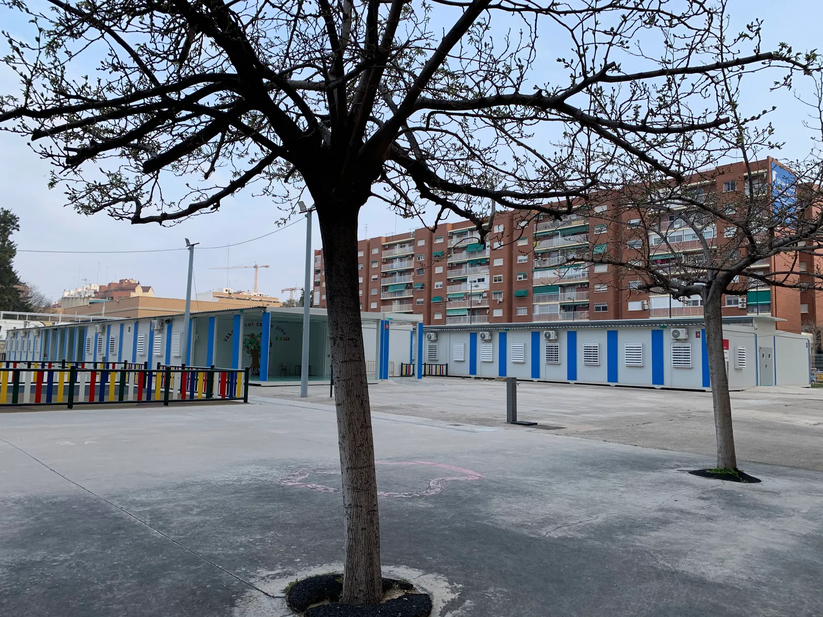 Empty primary school yard in Valencia with bare trees, low classroom blocks with blue stripes and a brick apartment block behind.